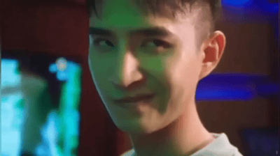 图片中是演员管云鹏的特写镜头，他嘴角微微上扬并向一侧歪斜，露出标志性的“歪嘴一笑”表情。背景为昏暗环境，带有蓝绿色灯光效果，整体氛围符合“龙王赘婿”系列广告中“身份揭露前的隐忍与即将逆袭的得意”场景。这一表情是2020年爆红的“龙王赘婿”文化现象的核心视觉符号，代表了网络爽文中“卑微赘婿逆袭打脸”的经典桥段。