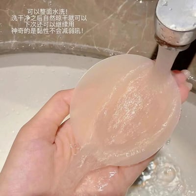 图片展示了一只手正在水龙头下冲洗一个透明的硅胶胸贴，水流正对着胸贴表面冲洗。背景是带有大理石纹理的洗手池台面，整体场景是在浴室环境中。左上角有产品说明文字，介绍该胸贴可以整面水洗，洗净晾干后可继续使用且黏性不会减弱。这是一个产品功能演示，展示了该硅胶胸贴的可重复使用特性。