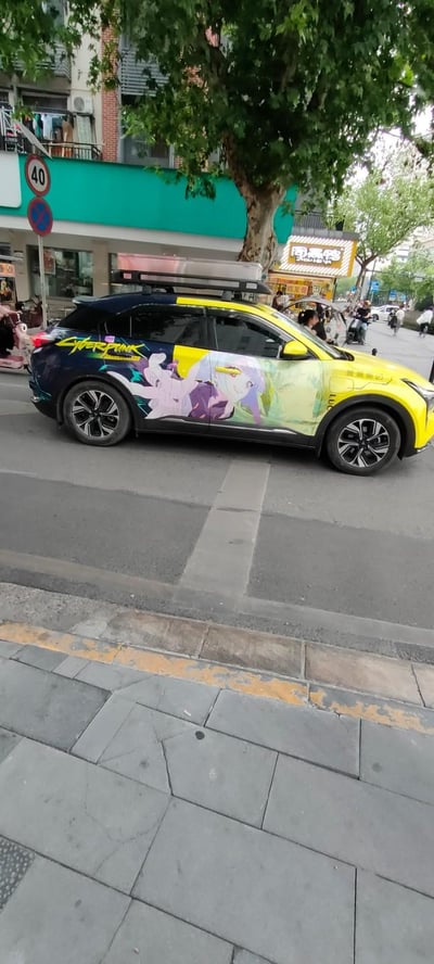 这是一张在城市街头实拍的痛车照片，车辆以黄蓝撞色为底色，车身侧面印有《赛博朋克：边缘行者》中的角色丽贝卡，还标注有"CYBERPUNK"字样，车顶加装了行李箱，背景可见临街商铺与行道树，是ACG爱好者结合汽车改装打造的个性化车辆，充满酷炫的个性风格。
