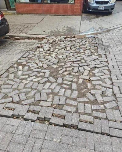 这是一张户外城市街景的实拍照片，画面核心是人行道上的灰色方形砖块出现了大面积的凸起、错位与变形：原本整齐排列的砖块被顶起，杂乱地堆叠在一起，形成了一块不规则的区域，与周围平整的人行道形成强烈反差，看起来十分怪异。背景中可以看到红砖建筑的墙角、停在路边的深色和银色汽车，是日常的城市街道场景，但路面的异常状况成为了视觉焦点，推测可能是地下管道故障、地基沉降或者冻胀等原因导致砖块拱起。