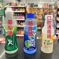 这是一张在超市场景下拍摄的P图改编梗图，画面里的三瓶常见饮料被添加了趣味文字：左侧雪碧瓶身标注“晕车喝”，中间脉动瓶身标注“感冒喝脉动”，右侧水溶C100瓶身标注“口腔溃疡喝”，通过谐音和场景关联，把普通饮料赋予了针对日常小病症的“专属功效”，用搞笑的方式调侃日常身体小不适。