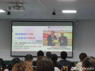 这是一张会议室现场拍摄的照片，画面中投影屏幕上方标注有“西南医科大学中西医结合学院附属医院”字样，屏幕左侧展示了该院院长杨思进的争议言论：“每天加班1小时，一生多活10多岁”，并通过偷换工作时长与自然寿命概念的计算方式来佐证该言论，将80年里每天加班1小时的总时长换算为相当于10年的工作时长，却偷换概念表述为“一生多活10多岁”；屏幕右侧是杨思进受聘担任岐伯生命健康文化研究院院长的现场照片，下方标注了相关受聘信息。画面下方可见多位参会人员背对镜头观看投影内容，照片右下角带有微博用户水印。该言论本质是混淆工作时间与生命时长的概念，属于美化加班、变相鼓励员工加班的不当宣传。