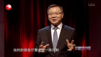 这是政论节目《这就是中国》的视频截图，学者张维为身着深色西装、白衬衫搭配花纹领带，戴着眼镜，面带亲和的微笑，双手做出手势正在进行演讲分享。画面下方配有字幕“当时的报告厅里出现一阵沉默”，背景是暗红色的幕布，整体场景正式，传递出演讲时的讲述氛围。