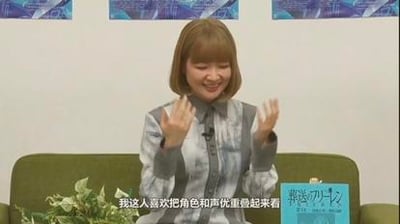 一位女性坐在绿色沙发上，身着蓝白格子衬衫，双手举起作比划状，面带灿烂微笑。背景墙上悬挂着蓝色海报，沙发右侧放置一本封面印有“声漫咖”字样的蓝色书籍。画面底部配有字幕“我这人喜欢把角色和声优重叠起来看”，整体场景呈现出轻松愉快的访谈氛围。