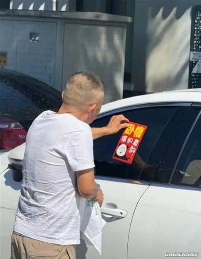 一位身穿白色T恤和卡其色短裤的中年男性，正站在一辆白色汽车旁，右手拿着一张红色贴纸，将其贴在汽车后窗上。贴纸上有黄色粗体文字“卧槽”和黑色文字“停的真好”，中间还配有一个卡通表情图案。背景中可见另一辆停放的汽车及灰色建筑物，整体场景捕捉了生活中用幽默方式吐槽停车情况的有趣瞬间。