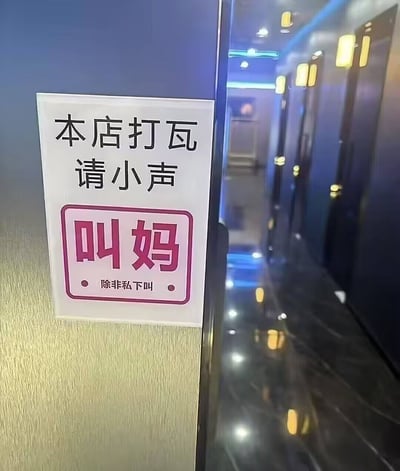图片展示了一处店铺走廊场景，走廊两侧有深色门扉，顶部装有暖黄色灯光，地面为反光材质，整体环境略显昏暗。左侧墙壁上挂着一块白色提示牌，上方用黑色字体写着“本店打瓦 请小声”，下方有一个粉色边框的矩形区域，内用红色大字写着“叫妈”，底部标注“除非私下叫”。标牌通过反差强烈的文字组合制造幽默效果，前半句“请小声”与后半句“叫妈”形成意外转折，暗示“打瓦”（可能为打牌、游戏等活动）时即使激动也需控制音量，避免大声“叫妈”，仅可私下表达，充满生活气息的趣味调侃。