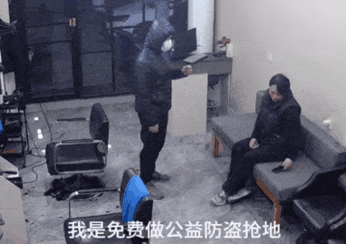 这是一段真人拍摄的动态GIF，场景设置在一个室内空间，地面散落着一些物品和椅子。一名身着深色带帽外套、佩戴口罩的男子，手持类似喷雾的物品，做出指向的动作，对着坐在沙发上的人说话；沙发上的人戴着耳机，神态平静地坐着。画面底部配有字幕“我是免费做公益防盗抢地”，利用表述的反差制造荒诞搞笑的效果——男子的对峙动作和“公益防盗抢”的说法形成强烈反差，让场景显得滑稽又离谱。