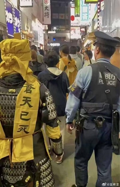 夜晚热闹的街道上，许多行人中混杂着几位身穿传统武士铠甲、披着写有“苍天已死”黄色绶带的人，旁边有现代警察同行，形成鲜明的古今对比场景。街道两旁挂满各式招牌，充满生活气息。
