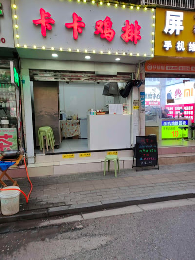 这是一张国内街边商铺的实拍图，主角是名为“卡卡鸡排”的小吃店：招牌采用红色立体发光字，外围环绕着一圈黄色小灯串，十分醒目。店铺为开放式操作空间，内部可见操作台、冰箱，还有堆叠的浅绿色塑料凳，店门口也摆放了同款凳子。店门口右侧立着一块手写黑板价目牌，标注着试营业优惠信息：黑椒鸡排16元、奥尔良鸡排16元，还有十元档位的餐品，购买鸡排赠送鸡肉洋葱圈，甘梅地瓜8元。
店铺右侧是一家手机维修店，招牌上有“快修猫”标识，还展示了华为、小米的品牌标志，提供手机维修、贴膜、回收等服务；左侧则是一家带有“茶”字的饮品店。整体呈现出日常、平实的城市街边商铺氛围，没有双关、谐音梗等特殊内容。