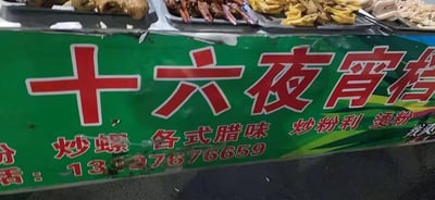 图片展示了一个街头夜宵档的招牌特写。招牌主体为绿色底色，上方用红色大字写着“十六夜宵档”，下方依次列出经营品类：“炒螺”“各式腊味”“炒粉利”“烫粉”，最底部标注了联系电话“13137676659”。招牌上方可见盛放食物的金属托盘，里面摆放着腊味等食材，整体呈现出浓郁的市井烟火气，是典型的城市街头小吃摊场景。
