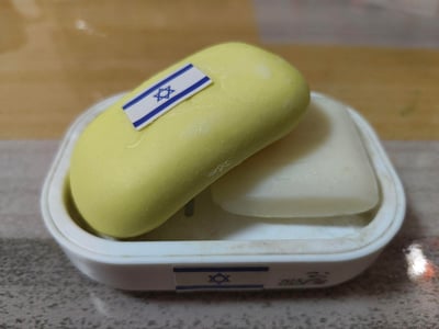 这是一张实拍的日常物品图，白色肥皂盒内放置着一块使用过的黄色肥皂和一块白色肥皂，黄色肥皂的表面粘贴了一张以色列国旗贴纸，肥皂盒的正面也贴有同款以色列国旗贴纸，皂盒右下角带有品牌标识。图片无双关、谐音梗或暗示类内容。