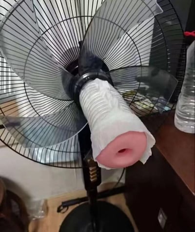 图片展示了一台黑色台式电风扇，扇叶呈透明状。风扇的中心电机部位被白色的卷纸筒包裹，卷纸筒末端连接着一个粉色的圆柱形物体，该物体顶端有一个圆形孔洞。风扇放置在木质桌面上，背景中可见一个透明的塑料水瓶。整体给人一种家庭DIY改造的感觉，可能是用户尝试制作某种特殊的送风装置，画面带有一定的搞笑和迷惑感。