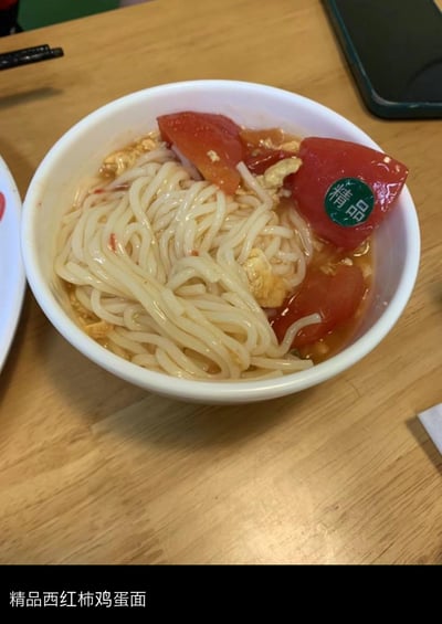 一张展示精品西红柿鸡蛋面的照片。白色碗中装着劲道的面条，浸泡在红色的番茄汤汁里，搭配大块番茄片和金黄的鸡蛋碎。其中一片番茄上贴着绿色的'精品'圆形贴纸，显得格外醒目。碗放置在木质餐桌上，旁边可见黑色手机、黑色笔和部分白色盘子，整体场景像是日常用餐时拍摄的真实画面。