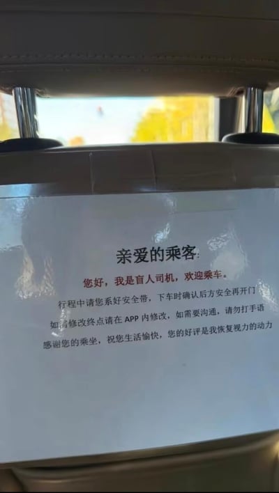 车内拍摄的乘客提示牌照片，内容为：'亲爱的乘客：您好，我是盲人司机，欢迎乘车。行程中请您系好安全带，下车时确认后方安全再开门。如需修改终点请在APP内修改，如需要沟通，请勿打手语。感谢您的乘坐，祝您生活愉快，您的好评是我恢复视力的动力。' 标语以荒诞幽默的方式虚构盲人司机身份，其中'好评是恢复视力的动力'带有明显的搞笑夸张效果，可能用于社交媒体分享博人一笑。