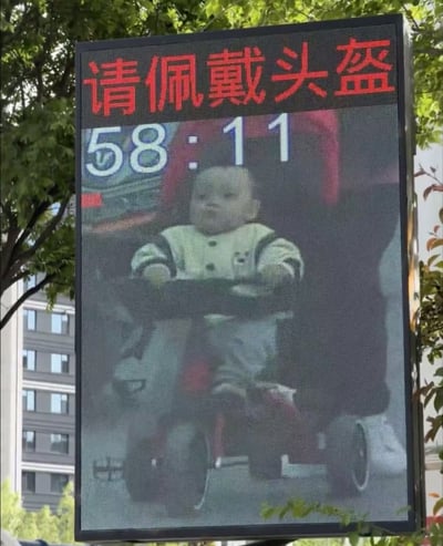 这是一张路人实拍的户外交通违法曝光电子屏的照片，电子屏顶部用醒目的红色大号字体标注了“请佩戴头盔”的电动车驾乘安全提醒，屏幕下半部分展示了抓拍的违法画面：一名幼儿坐在电动车的前置儿童座椅上，全程未佩戴安全头盔，画面上方还显示有白色数字“58:11”，电子屏的后方背景可以看到翠绿的行道树和高层居民楼。该设备本是用于公示驾乘电动车未佩戴头盔的成年违法人员，本次抓拍到的违法主体为低龄幼儿，内容具有反差趣味。