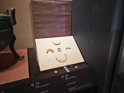 博物馆展柜中展示着战国时期的玉璜文物，出土于漳州市牛山古墓群。展品被放置在白色基座上，周围有中英文标签说明。左侧可见一件青铜器皿，整体场景为博物馆内的文物陈列，展现了古代玉器文化。