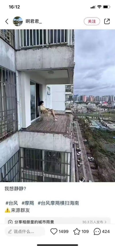 这是一张带有视觉错位效果的实拍图，在台风“摩羯”横扫海南后的城市背景中，一位女士淡定地坐在高层住宅的露台躺椅上，姿态放松悠闲。背景里能看到被台风影响的城市景象：路边车辆倾倒、天空阴沉，与女士的淡定状态形成强烈反差。配文“我想静静？”，结合场景既表达了想安静放空的心情，也用反差感调侃了台风后的氛围。露台的建筑结构在拍摄角度下看起来像是“悬空”，制造出惊险又淡定的视觉效果。