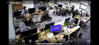 这是一张办公室监控的视频截图，画面呈现开放式办公区场景，多个工位上摆放着电脑、办公桌椅与各类办公杂物，数位员工正在工位上忙碌或休闲。前景处一位戴帽子的女生的电脑屏幕上，显示着“看你妈b”的粗俗恶搞文字，在严肃的监控视角下形成强烈反差，充满搞笑感，是员工在监控视角下的职场整活行为。