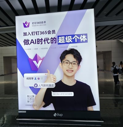 地铁站内的大型竖版广告牌，主体为钉钉365会员推广内容。画面中央是人气数码博主何同学，他穿着黑色T恤，面带微笑并竖起右手大拇指，胸前配有文字气泡标注“我已加入钉钉365会员”。广告牌顶部左侧有紫色皇冠造型的钉钉365会员logo及标语“有创造力的人用钉钉365”，主宣传语“加入钉钉365会员 做AI时代的超级个体”以蓝紫色渐变背景突出显示，下方罗列个人AI助理、AI搜索、AI自动回复等六项会员权益。左下角设有二维码，底部中央有“duup”标识。背景为公共交通枢纽的走廊场景，有行人模糊经过。