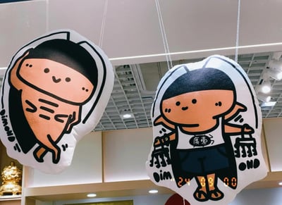 图片中展示了两个悬挂的卡通蟑螂毛绒玩具，基于2024年末爆红的'广式双马尾'梗创作。左侧玩具呈橙色，有着黑色的'双马尾'触角，面带微笑，身体圆润；右侧玩具穿着印有'广东'字样的白色背心和深色短裤，手持购物袋，脚下有'24:00:00'的数字显示，整体造型可爱，将蟑螂的触角拟人化为双马尾，体现了网友用幽默化解生活困扰的乐观精神。