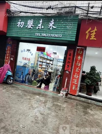 这是一张充满生活气息的街边店铺实拍图，主体是名为“初婴未来”的母婴用品店。店铺招牌为绿色底白色字体，下方标注联系电话18972703613。玻璃门敞开着，店内货架上摆满了各类母婴商品，门口的玻璃上贴着“欢迎光临”的字样。店铺两侧张贴着红色春联，右侧为“好年好景好運氣”，左侧为“喜迎新春開”（部分文字被遮挡），左侧门边还停放着一辆蓝粉配色的电动车。一位穿着黑色衣服的人坐在店铺门口的紫色小凳子上，双腿交叠，姿态放松。地面看起来潮湿，似乎刚下过雨，整体场景展现了普通街边店铺的日常样貌。