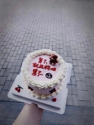 这是一张拍摄于室外的蛋糕图片。蛋糕表面写着“第了，做这些烂事累了”，配有一个熊造型装饰，旁边画了一个表情汗颜的小人头，蛋糕边缘点缀着红樱桃。整体画面带有无奈、网友自嘲的情绪，是近年流行的“摆烂”“丧文化”释压梗，用以表达对生活琐事、压力感到疲惫，调侃且自嘲地展示“累了不干了”的状态。本图通过庆祝场景与丧气内容反差形成幽默梗效果。

特殊内容说明：
- “第了”为网友打错字的“累了”，这种错字加剧了自嘲氛围，充满网络化幽默感。
- 蛋糕搭配消极短语表达反向庆祝，成为独特网络段子模式。