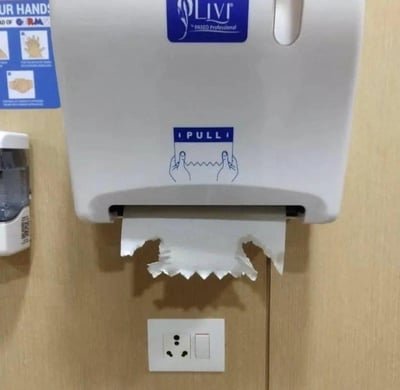 这是一张真实场景的实拍图，画面中是安装在墙面上的LIVI品牌擦手纸盒，纸盒上印有「PULL」的取纸提示，示意使用者捏住纸巾锯齿部分拉取。而纸盒下方露出的纸巾被撕出了和正下方插座完全匹配的缺口，仿佛是为了避开插座特意撕成这个形状，形成了极具趣味性的巧合，让人忍俊不禁。