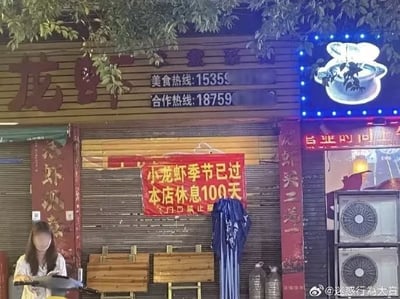 夜晚的龙虾店门头实拍图，招牌上方有“龙虾”大字及模糊的“美食热线:1535C”“合作热线:18759”字样。店门正上方悬挂红色横幅，醒目写着“小龙虾季节已过 本店休息100天”。一名面部被模糊处理的女子坐在店前木质桌椅旁，手中拿着锅具。店铺右侧有蓝色LED灯装饰的锅形发光招牌，上方有树叶遮挡，整体呈现夜间街边店铺的真实场景。