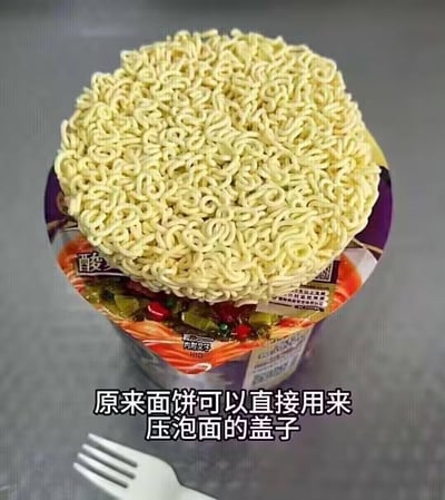 这是一张展示生活小技巧的实拍图：将未冲泡的泡面面饼直接放置在打开的桶装泡面盖子上，用来压住盖子，替代叉子压盖的常规方式。图中的桶装泡面带有“酸”字标识，推测是酸辣口味，面饼完全覆盖住盖子，下方可见泡面的料包和包装上的“内附叉子”字样，图片底部配文“原来面饼可以直接用来压泡面的盖子”，这个用法新奇又实用，自带搞笑感。