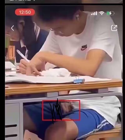 这是一张课堂场景的屏幕截图，一名穿着白色耐克T恤的学生正低头专注地在本子上书写作业。画面下方的蓝色衣物区域因视觉错位，看起来像是有一只小动物趴在腿上，实际是衣物褶皱与视角共同造成的错觉，制造出滑稽搞笑的意外效果。
这是典型的视觉错位类趣味内容，利用视角偏差营造出反差感。
