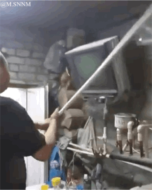 这是一段趣味动态GIF，画面中一名男子身处堆满工具和杂物的杂乱房间内，手持长杆做出挥打动作。借助视觉错位效果，长杆看起来像是正击打老式电视机的屏幕，仿佛男子在和屏幕里的内容互动，实则杆子从电视机旁穿过，制造出滑稽的视觉错觉，充满搞笑感。