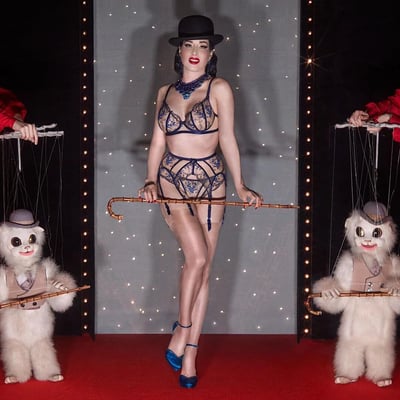 图片中展示了一位身着蓝色蕾丝内衣的女性站在舞台中央，她佩戴着黑色礼帽和蓝色宝石项链，手持一根长拐杖，姿态优雅自信。她的两侧各有一个白色提线木偶，这些木偶穿着类似的服装并同样手持小拐杖，由幕后人员操控着提线。背景是灰色幕布，点缀着星星状的灯光，两侧有串灯装饰，脚下是红色地毯，整体营造出复古华丽的舞台氛围。