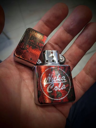 一只手握着一个打开的Zippo风格打火机，打火机具有红黑相间的大理石花纹外观，正面印有辐射游戏系列中的Nuka Cola标志。打火机外壳呈现出金属质感，开盖状态展示了内部的打火轮和防风孔结构。