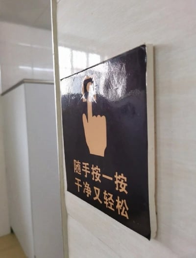 这是一张实拍的公共卫生间场景照片，墙面上张贴着一块黑底浅棕色文字与图案的冲水提示牌，提示牌原本设计为伸出食指按下冲水按钮的手势，搭配提示语“随手按一按，干净又轻松”，用于提示使用者便后冲水保持清洁。但提示牌上代表冲水按钮的图案部分出现磨损脱落，恰好让原本的食指按按钮的手势视觉上变成了竖起中指的样子，形成意料之外的搞笑恶搞效果，背景可以看到卫生间的隔间挡板。