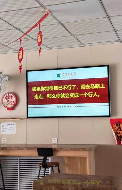 这张实拍静态图拍摄于室内公共区域，墙面悬挂的石河子大学电子显示屏上展示了一则趣味标语：红底黄字写着“如果你觉得自己不行了，就去马路上走走，那么你就会变成一个行人”，利用“行人”的双关含义（一是指在道路上行走的人，二是指“能干、可以的人”）制造谐音梗，达到搞笑又励志的效果，屏幕底部还印有石河子大学校训“明德正行 博学求真”。画面中还能看到天花板悬挂的新年福字挂饰、墙面的2025年贺年装饰、立式风扇、吧台座椅、“前程似锦”的红色新年装饰，整体充满生活气息，是学校发布的趣味治愈类宣传内容。