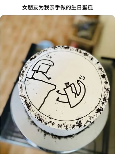图片展示了一个由女朋友亲手制作的生日蛋糕。蛋糕整体为圆形，表面覆盖着白色奶油，边缘撒有黑色碎屑（疑似奥利奥碎）。蛋糕顶部用黑色线条手绘了两个简笔画人物：左侧人物旁标注“24”，呈现出坐在台阶上的姿态；右侧人物旁标注“23”，姿态像是在鞠躬或递东西。蛋糕上方配有文字“女朋友为我亲手做的生日蛋糕”，整体画面温馨中带着幽默，手绘图案的“灵魂画手”风格突出了手工制作的真诚与可爱。
