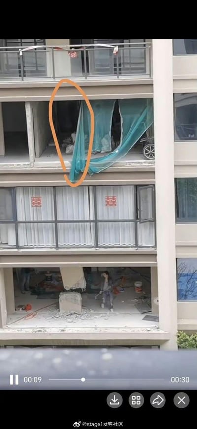 这是一段竖屏视频的截图，展示了一栋高层住宅的多户状态：最下方楼层室内有大量拆改产生的建筑垃圾，一名人员身处其中，看起来有些慌乱；中间楼层的窗户挂着窗帘，玻璃上贴有两个红色“囍”字，推测是刚布置完的婚房；上方被橙色圈出的楼层，楼板出现坍塌破损，绿色防护布被砸出大洞，内部可见堆积的建筑垃圾。整个场景显示该住宅存在疑似违规拆改建筑结构的行为，进而引发上层楼板坍塌，存在极大的建筑安全隐患。