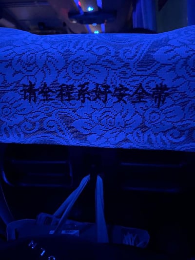 这是一张在客运客车内部拍摄的竖版照片，画面主体是前排座椅的靠背罩，靠背罩带有复古花纹样式，上面印有深色的提示文字“请全程系好安全带”。画面下方可以看到座椅安全带的部分结构，整体环境光线偏暗，被蓝色冷色调光线笼罩，推测是在夜间或者光线不足的车内环境下拍摄。该标语是客运车辆的安全提示内容，用于提醒乘客在乘车全程佩戴安全带，没有双关、谐音梗或其他暗示内容。