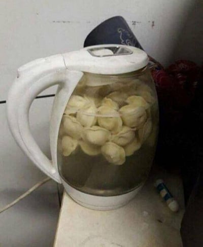 图中展示了一个白色的玻璃电热水壶，壶内盛放着水和多颗类似俄式水饺（馄饨）的速冻面食，电热水壶被当作煮锅来使用，放置在一个简易的台面上，旁边还有一个小的圆柱形物件。这是一种非常规的烹饪方式，用原本用来烧开水的电热水壶来煮食物，显得十分奇葩、搞笑，充满了生活里的硬核懒人操作感。
