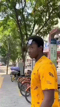 这是一段竖屏GIF动态图，画面中一位身着黄色带图案上衣的黑人小哥站立在城市街边，背景可见行道树、停放的共享单车以及街边商铺。由于视觉错位的巧合，后方的自行车轮与小哥的额头精准重合，看起来仿佛小哥的额头上有一个圆形印记，制造出十分有趣的视觉错觉效果，让人忍俊不禁。