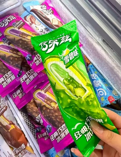 这张图拍摄于商超的冰淇淋冷柜区域，一只手举着一包全新的亮绿色包装巧乐兹冰淇淋，这款是新推出的雪碧味巧乐兹经典巧恋果冰淇淋，包装上印有对应青绿色雪糕的剖面示意图；背景的冷柜里还摆放着多包紫色包装的巧乐兹蓝莓味冰淇淋、其他口味的巧乐兹产品以及蓝色包装的可爱多冰淇淋，整体是消费者实拍分享市面新奇雪糕口味的场景。