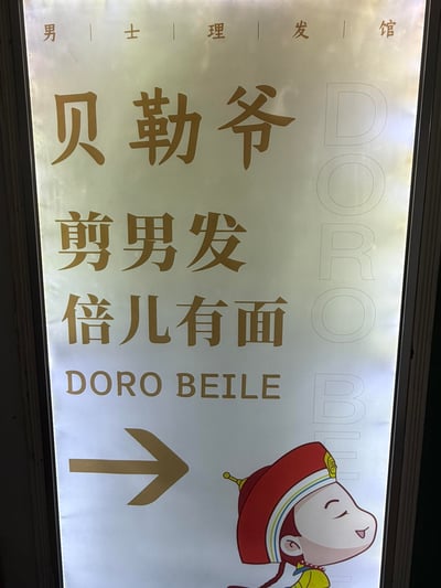 这是一块竖版的男士理发馆指示灯牌，顶部以竖线分隔标注「男士理发馆」，主视觉为大号金色字体的「贝勒爷」，下方搭配宣传语「剪男发 倍儿有面」，还有对应的英文「DORO BEILE」；灯牌右侧有浅灰色竖排的「DORO BEI」字样，右下角是闭着眼睛、面带笑意的卡通贝勒形象，同时配有一个指向右侧的箭头，用来指引店铺方向。
其中「倍儿有面」是北京方言，意为很有面子，结合「贝勒爷」的京味儿IP定位，传递出到店理发能让顾客获得体面感的宣传点。