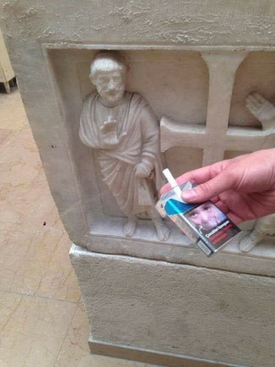 一张创意摄影作品，展示了一座古老的石雕人物像与现代物品的趣味互动。画面中，一座石制浮雕上的古代人物身着长袍，右手自然抬起，手掌朝前，仿佛在礼貌地索要或接住什么东西。一只现代人的手正拿着一盒"Parlament"香烟，将烟盒递向石雕人物伸出的手，巧妙地利用视觉错位营造出古人也想抽烟的幽默效果。这种古今对比和拟人化的创意，让严肃的历史文物瞬间变得生动有趣，配合标题"还怪有礼貌的👍"，形成了强烈的喜剧效果。