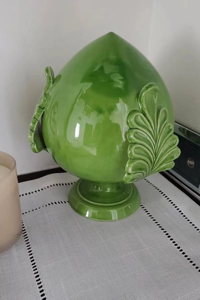 这是一张展示绿色陶瓷花瓶的照片。花瓶采用寿桃造型，整体呈圆润的桃形，表面光滑有光泽，呈现出明亮的绿色。花瓶两侧装饰有对称的叶子形状纹饰，底部有圆形底座。花瓶被放置在铺有白色带黑色缝线桌布的桌子上，背景中可见一个白色蜡烛和一个带有银色旋钮的黑色设备。