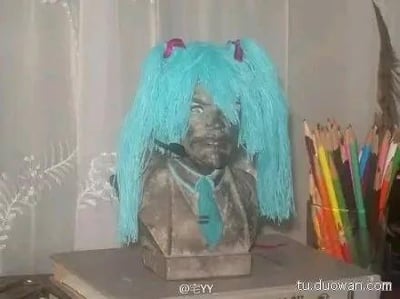 图中展示了一个被趣味改造的男性半身雕像，原本表情严肃庄重的雕像被戴上了虚拟偶像初音未来标志性的蓝绿色双马尾假发，还配上了同色系的领带，严肃的雕像形象和二次元偶像的元素形成强烈反差，制造出滑稽搞笑的效果，是ACG爱好者对普通物品的趣味玩梗改造。