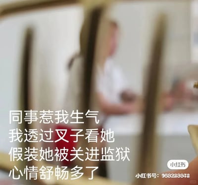 这是一张职场场景的实拍静态图，前景是叉子的齿状部分，透过叉子的齿看向背景，能看到模糊的职场同事身影。图中搭配了幽默的文字配文，讲述了作者被同事惹生气后，通过透过叉子的齿看同事，将叉子的齿想象成监狱栏杆，假装同事被关进监狱的方式，来疏解自己的负面情绪，最终心情舒畅的趣事。整张图用轻松搞笑的方式，展现了职场人应对职场小矛盾、疏解情绪的创意小妙招，容易引发职场群体的共鸣。