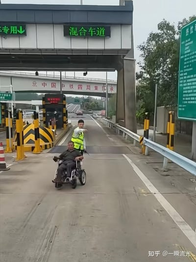 这张图片拍摄于高速公路收费站的混合车道处，一位男士正乘坐电动轮椅沿着收费车道向前行驶，后方的收费站工作人员身穿荧光反光背心，伸出手臂做出引导的动作，似乎在指挥轮椅的通行路线。背景中可以看到上方桥梁上的“中国中铁四局集团承”字样，收费站上方的电子标识牌显示“混合车道”和“专用↓”的指引，右侧还有一块绿色的服务区信息指示牌。整个场景比较少见，因为电动轮椅上高速的情况并不普遍，显得有些出人意料。