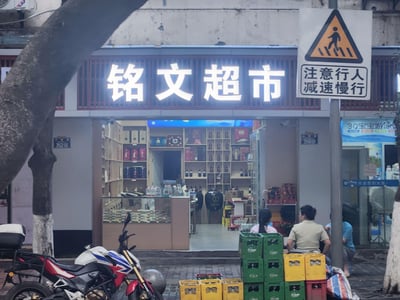 这是一张城市街头的实拍照片，画面核心是名为“铭文超市”的社区超市，店铺招牌为发光白色字体，店内货架上陈列着烟酒类商品，店门两侧门牌号分别为352和350。超市外的人行道上，三位市民正坐着休息，旁边堆叠着黄绿配色的饮料箱；左侧停放着一辆红蓝白三色的摩托车，画面右侧有一个“注意行人 减速慢行”的交通警示标志，旁边还有一家“3Q宝宝游泳”的店铺，整体呈现出充满生活气息的社区街边场景，没有双关、谐音梗等特殊内容。