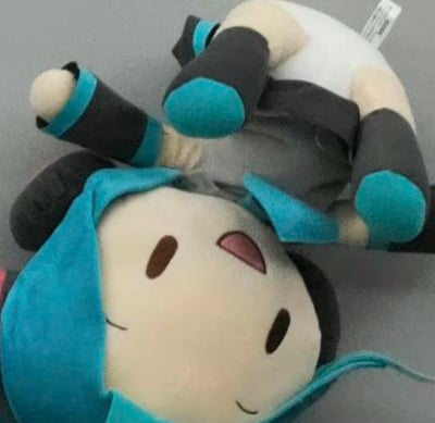 这张图展示了初音未来的毛绒玩偶，通过刻意的摆放制造出趣味视觉错位：玩偶的头部平放在下方，面部带着放松的微笑，而玩偶的身体部分被放置在头部上方，从拍摄角度看仿佛身体和头部“分离”且压在头部之上，形成滑稽又呆萌的画面效果，充满了生活化的趣味感。