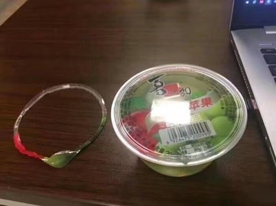 图中展示了放置在木质桌面上的一盒喜之郎苹果味果冻，旁边是被完整扯下来的果冻盒封盖拉环，这个拉环因为被整个撕下，形状类似一个透明手环，是日常里的趣味小意外场景，没有谐音梗或双关含义。